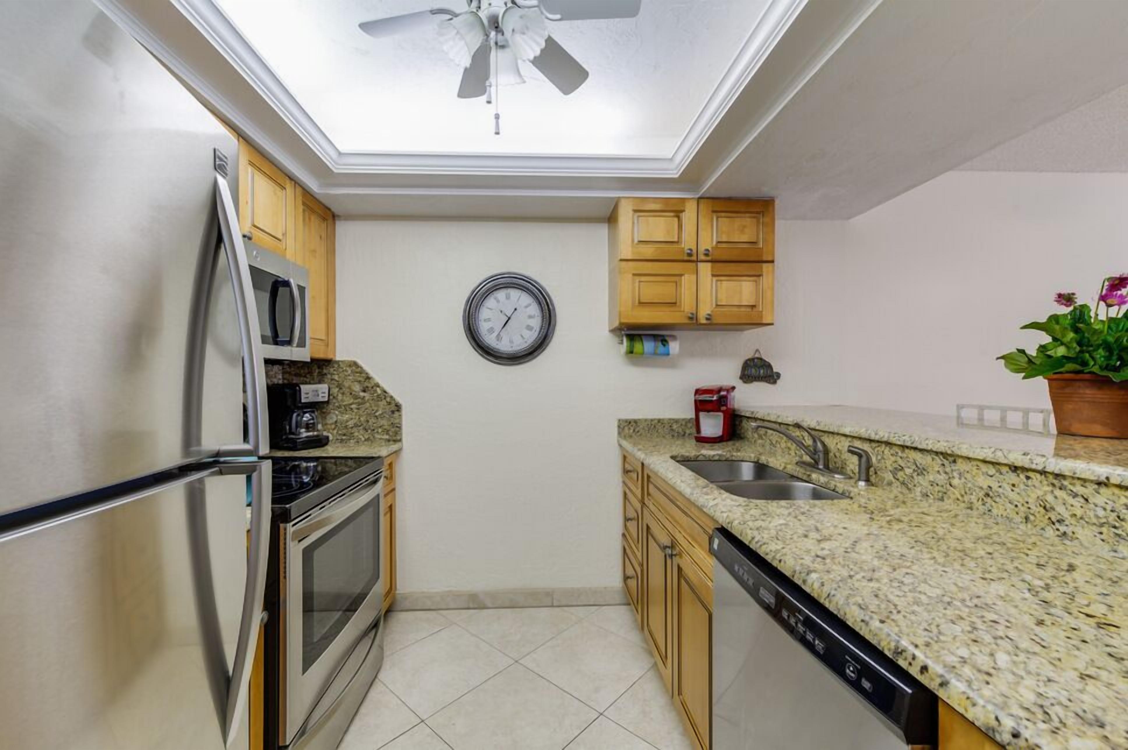 A compact kitchen with a refrigerator, oven, microwave, and dishwasher. A ceiling fan is visible above, and a clock hangs on the wall.