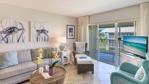 A modern living room with a sofa, armchair, and a television displaying a beach scene. There are framed artworks on the wall and a balcony with a view of the ocean. - Sanibel Siesta on the Beach (Sanibel)