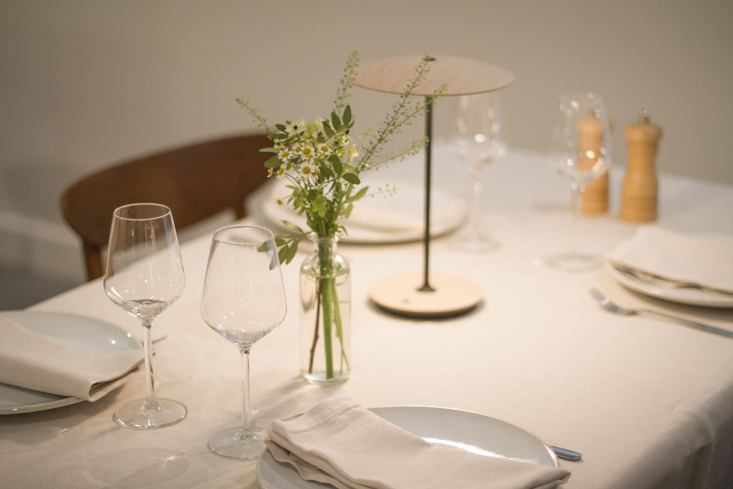 Ein Esstisch ist gedeckt mit weißen Tellern, Weingläsern, einer Blumenvase und einer kleinen Tischlampe.