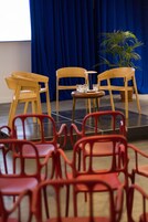 Eine Bühne mit blauem Vorhang, hölzernen Stühlen und einem kleinen Tisch mit Gläsern.