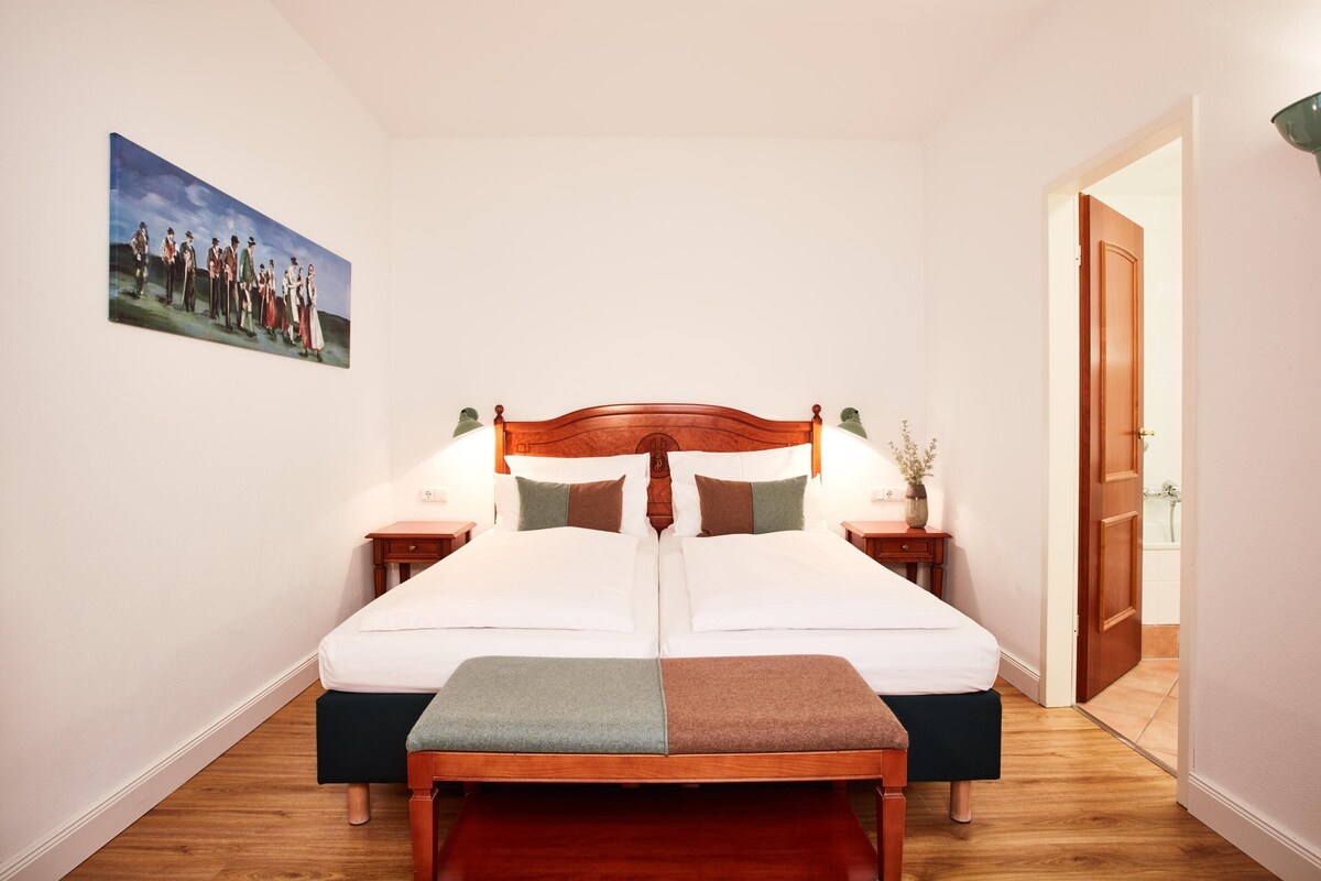 A double bed with a wooden headboard, two bedside tables with lamps, a painting on the wall, and a wooden floor.