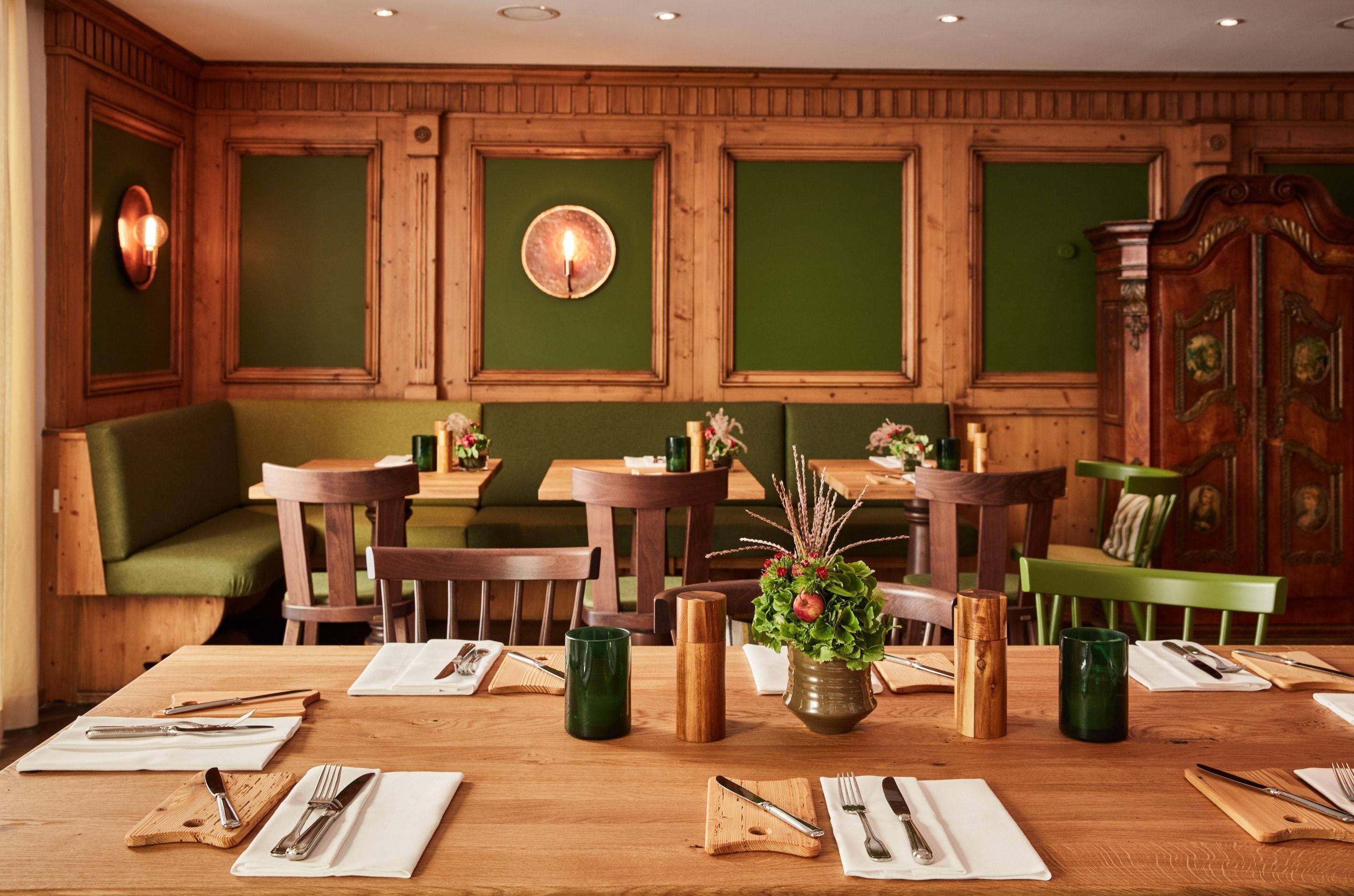 A dining area with wooden tables, green upholstered chairs, and a green wall with wooden paneling.