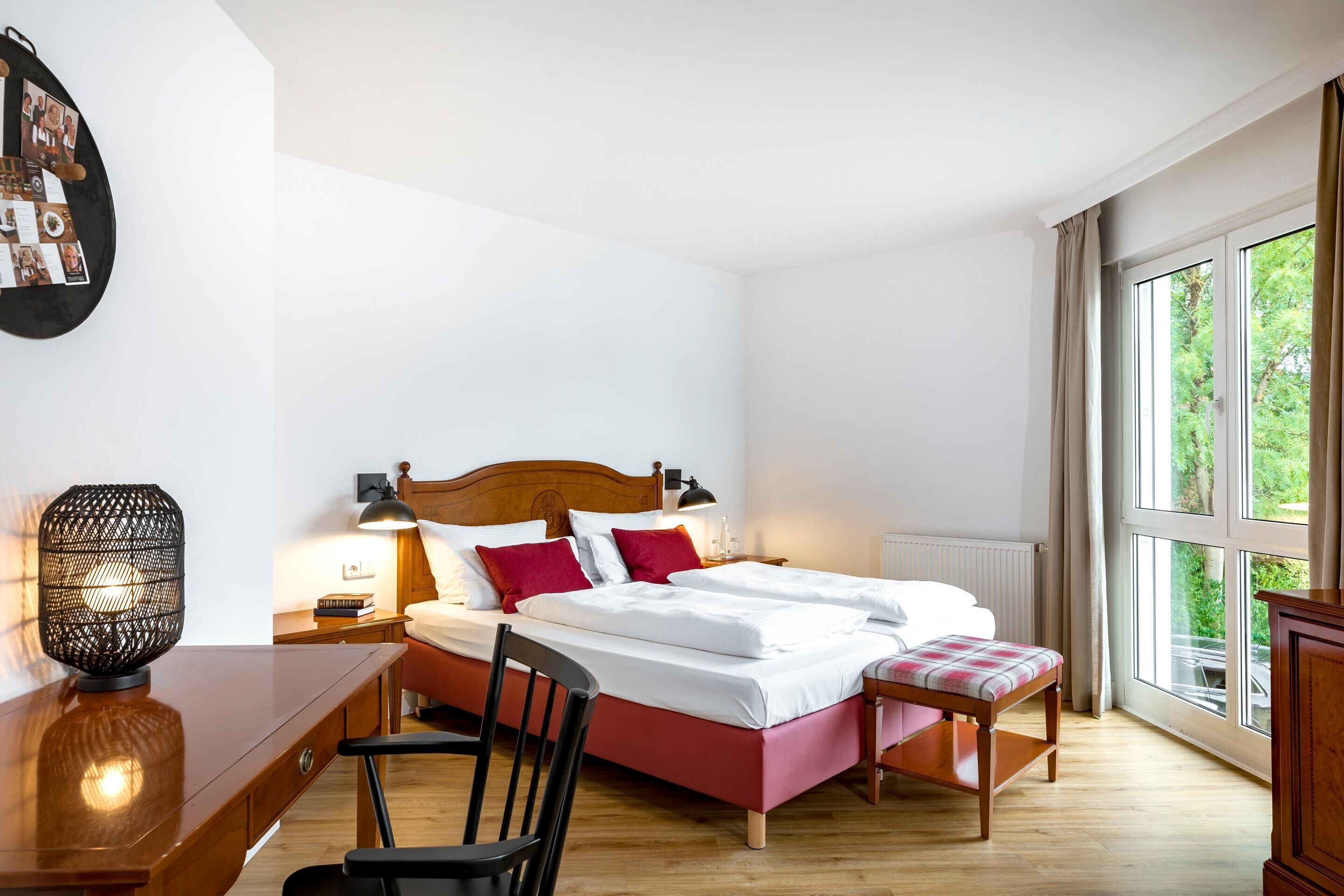 A bedroom with a wooden bed, a desk, a chair, a lamp, and a window with a view of greenery.