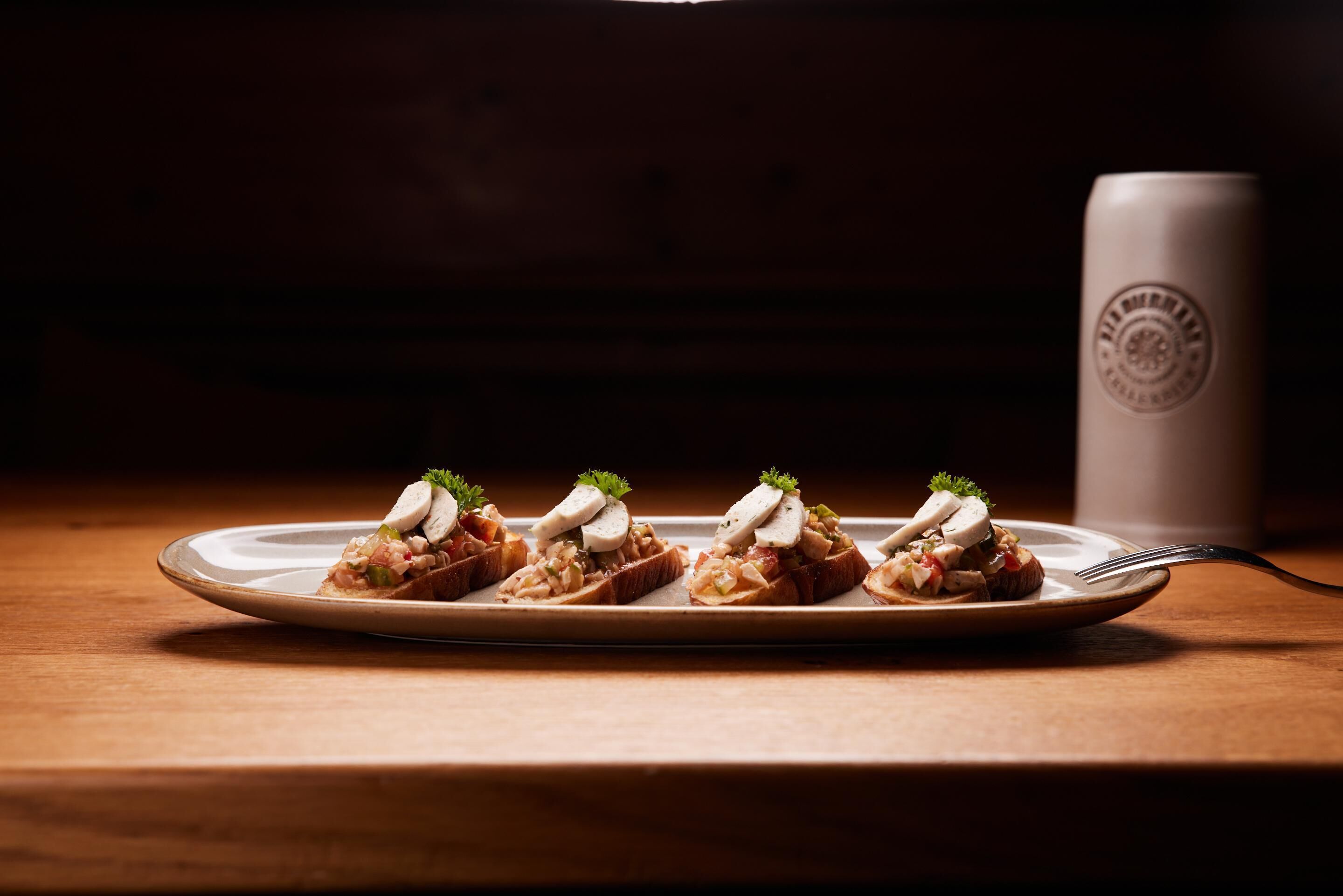 A plate with four pieces of food topped with cream and garnished with parsley, placed on a wooden table with a cup in the background.