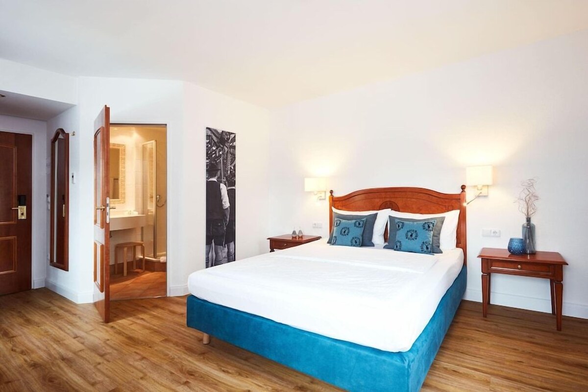 A bedroom with a large bed, wooden headboard, two bedside tables with lamps, and a bathroom visible through an open door.