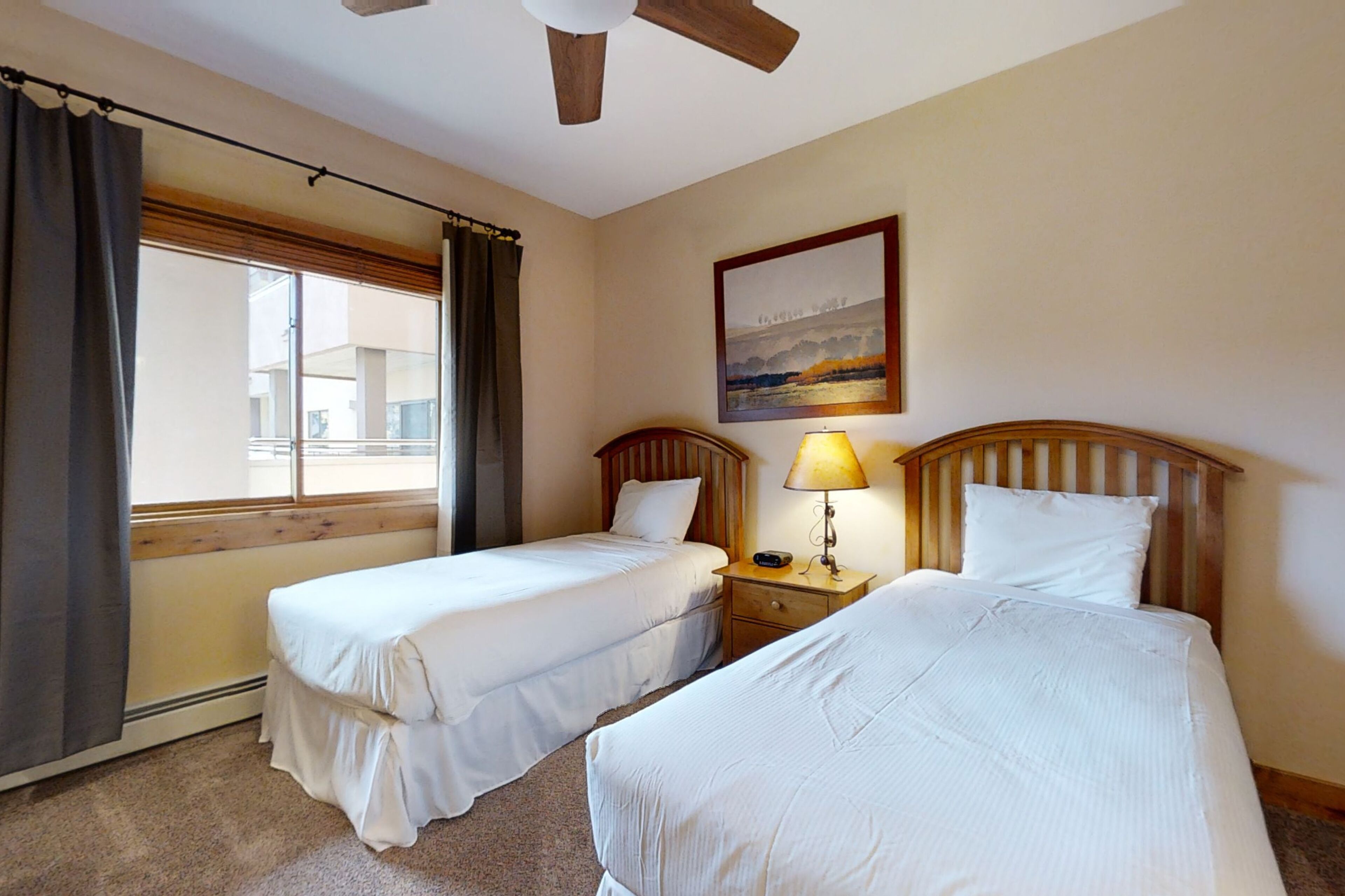 A hotel room with two beds, a ceiling fan, a window with curtains, a bedside table with a lamp, and a painting on the wall.