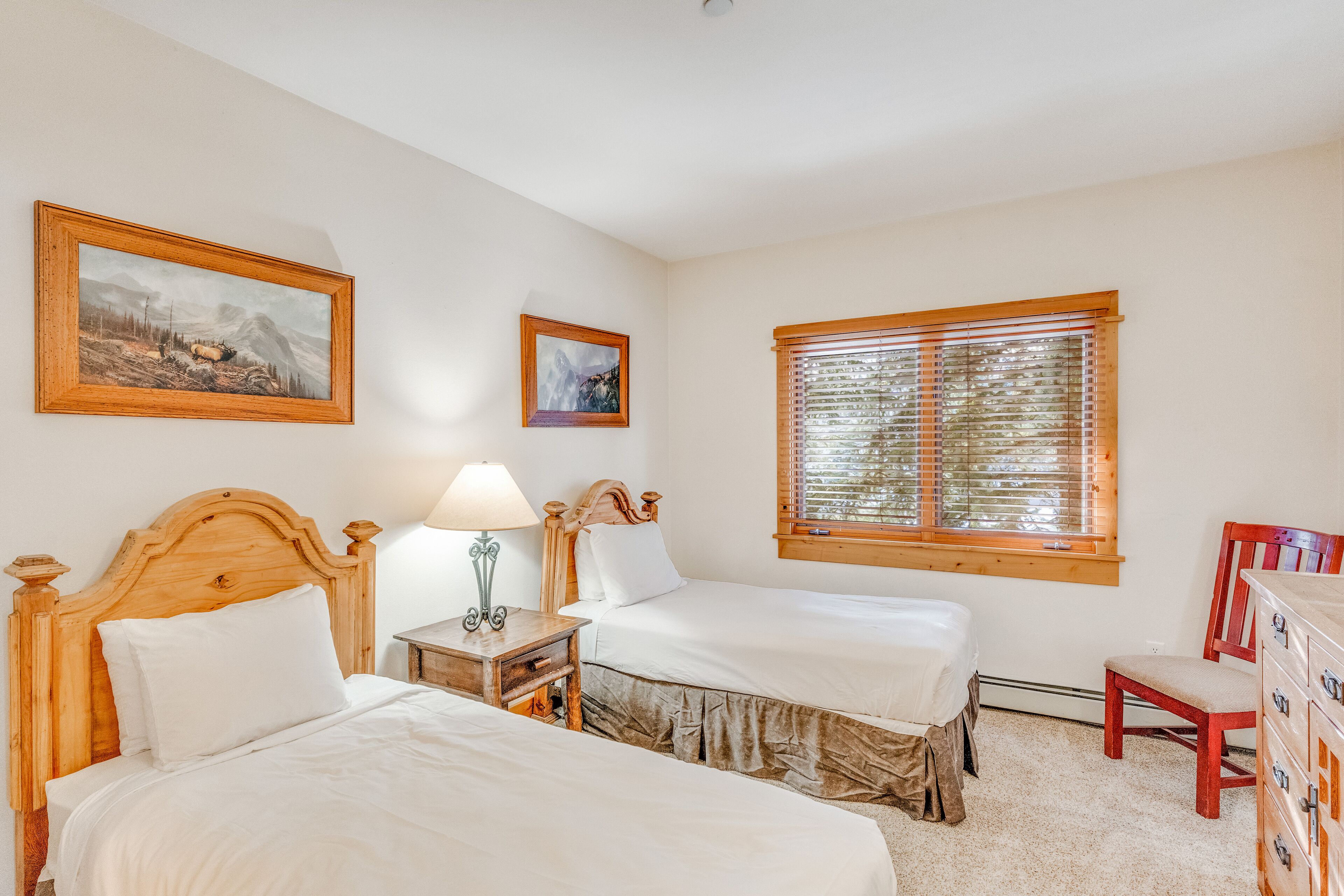 Une chambre avec deux lits, une tête de lit en bois, une table de chevet avec une lampe, une commode et une fenêtre avec des stores.