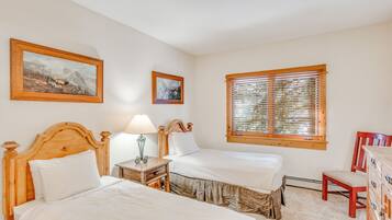 Une chambre avec deux lits, une tête de lit en bois, une table de chevet avec une lampe, une commode et une fenêtre avec des stores.