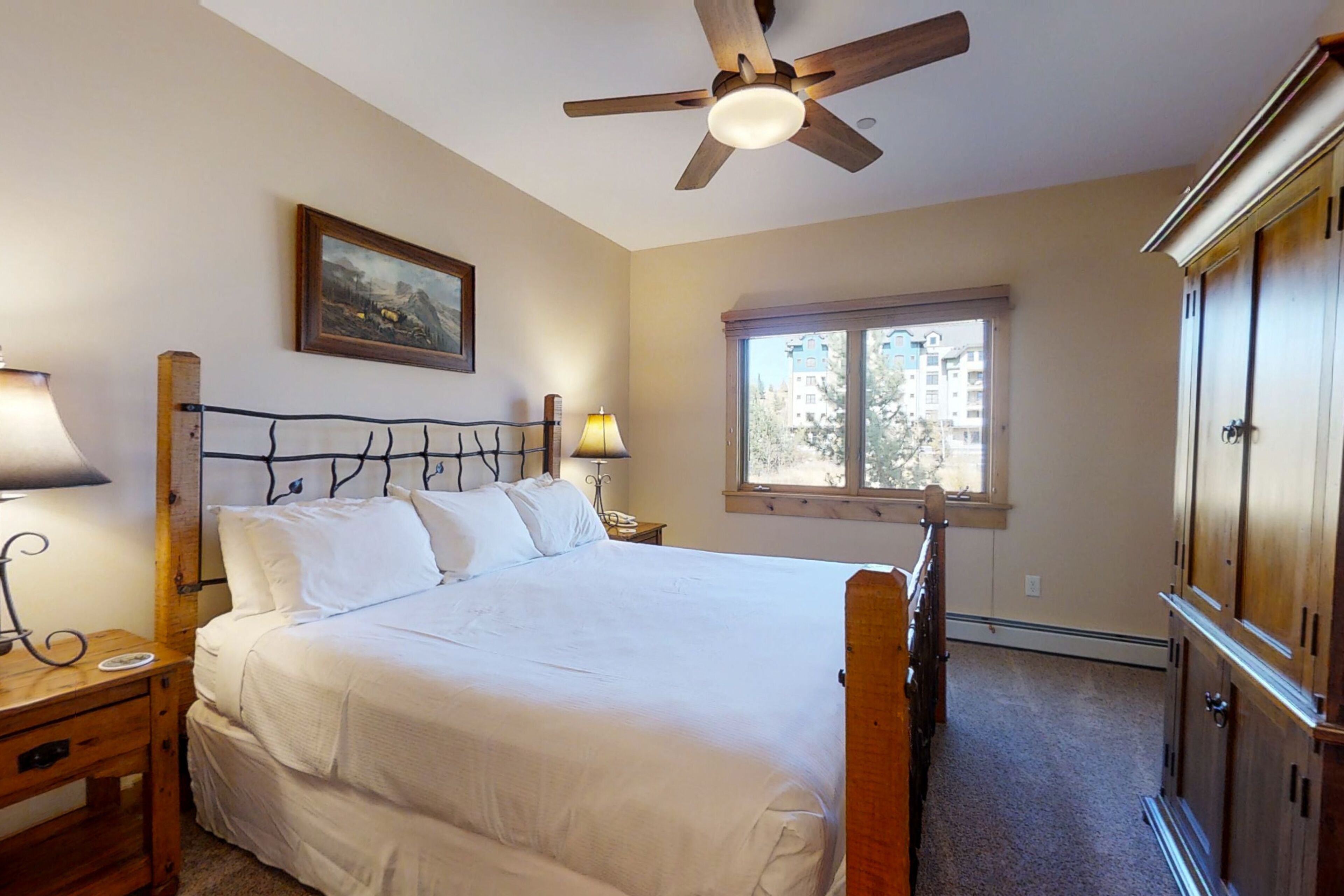 A bedroom with a bed, a ceiling fan, a window with a view of buildings, and a wooden wardrobe.