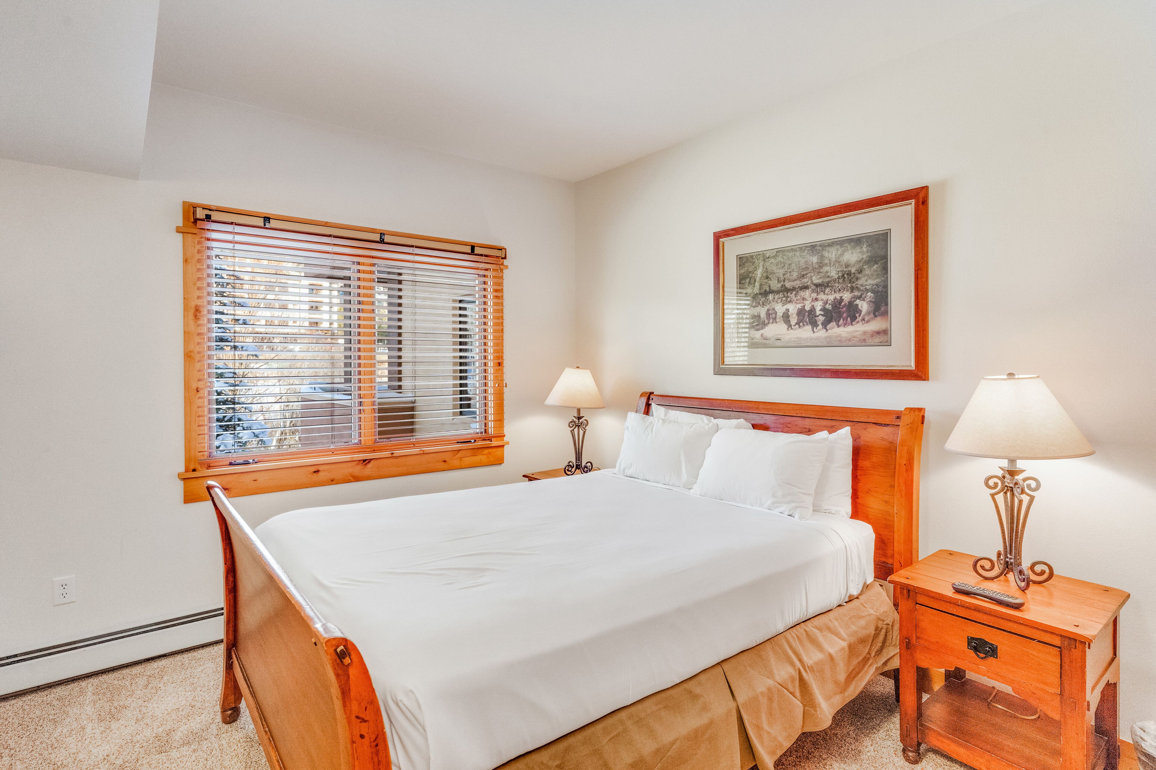 A neatly made bed with a wooden headboard, two bedside tables with lamps, a framed picture on the wall, and a window with blinds.