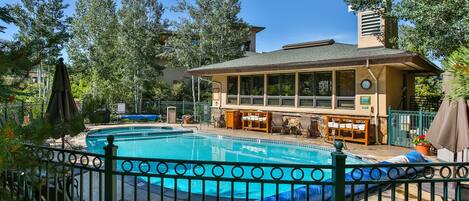 A swimming pool area with a fenced deck, lounge chairs, and a building with large windows and a chimney.