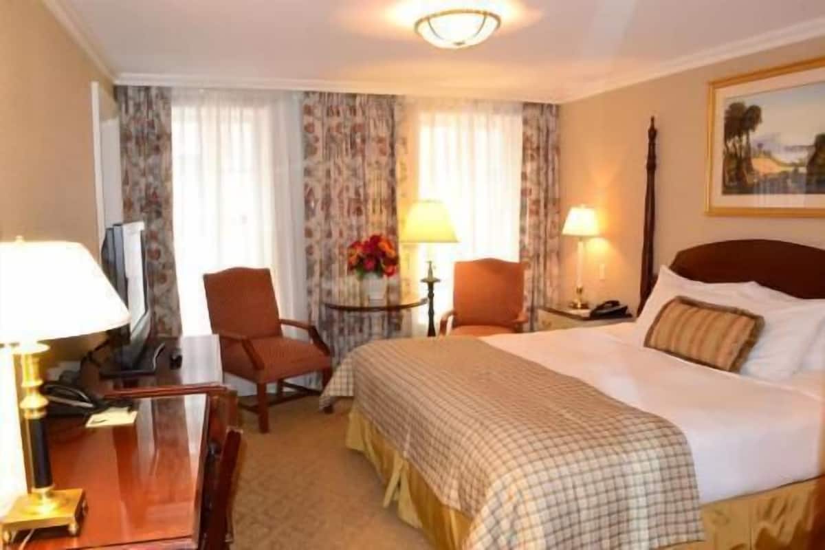 A hotel room with a bed, two chairs, a desk, a lamp, and a painting on the wall.