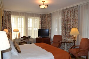 A hotel room with a large bed, two chairs, a television, and a desk with a lamp.
