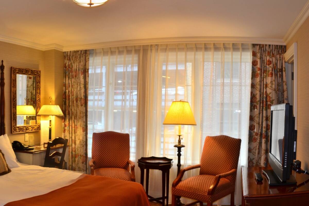 A hotel room with a large bed, two chairs, a desk with a lamp, a television, and a window with curtains.