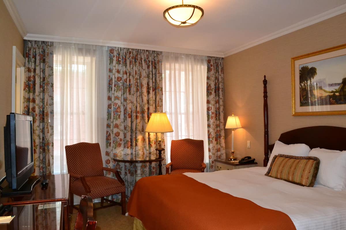 A hotel room with a large bed, two chairs, a desk with a television, and two lamps.