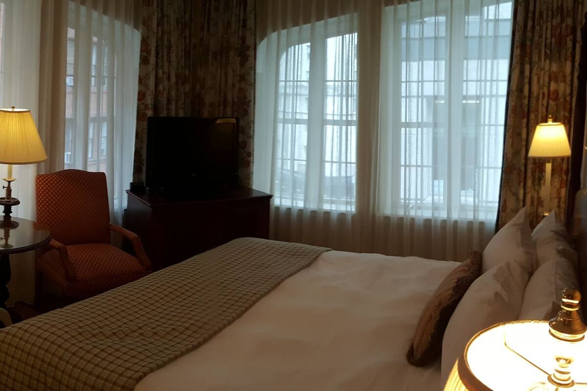A hotel room with a bed, two chairs, a small table, a TV, and two lamps.