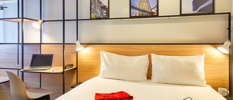 A hotel room with a bed, a desk with a laptop, a chair, and a shelf with pictures.