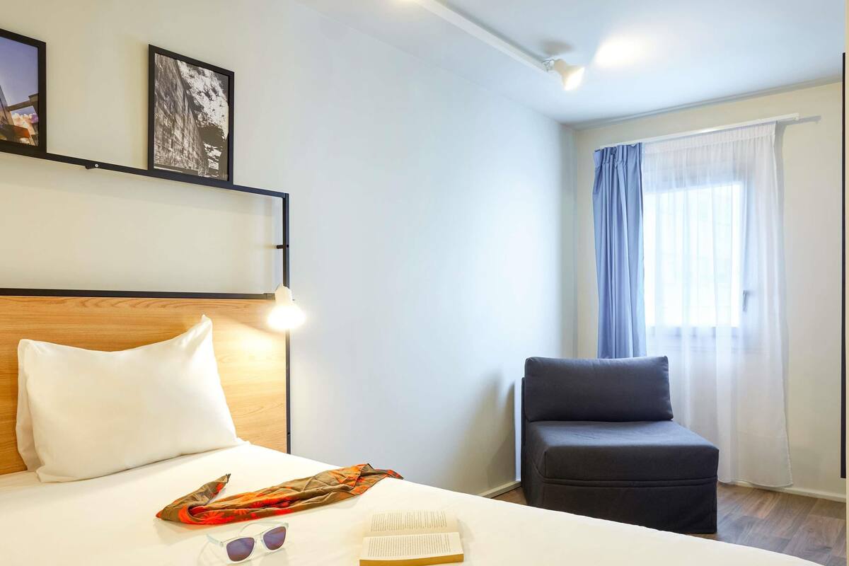 Ein Hotelzimmer mit Bett, Stuhl, Fenster mit Vorhängen und zwei gerahmten Bildern an der Wand.