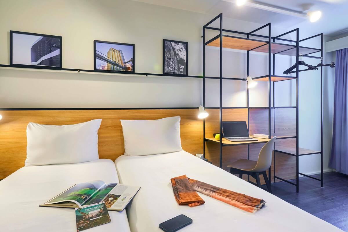 Ein Hotelzimmer mit einem Bett, einem Schreibtisch und einem Stuhl. An den Wänden hängen Bilder und auf dem Schreibtisch steht ein Laptop.