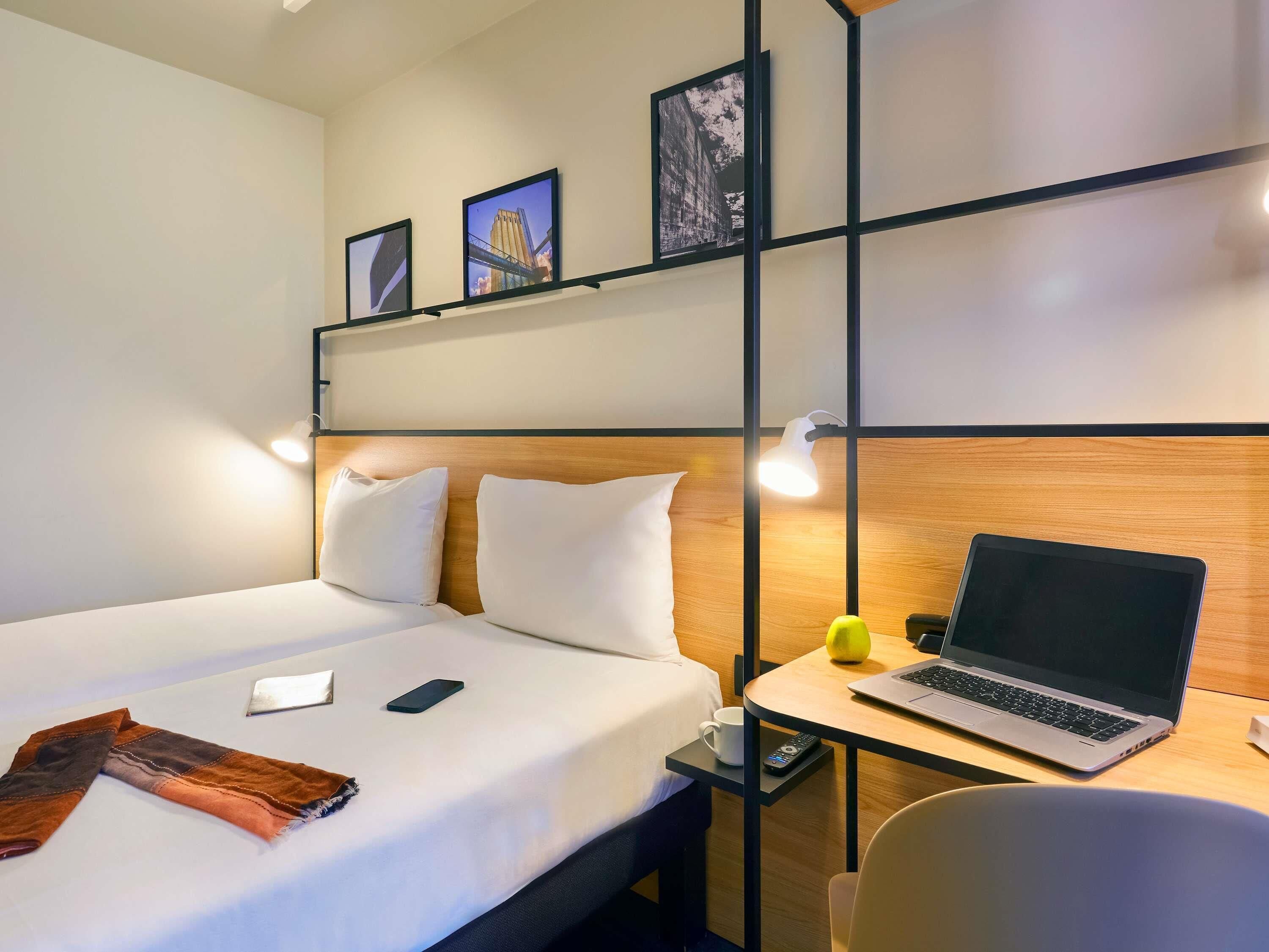 Una habitación de hotel con una cama, un escritorio con una computadora portátil, una silla y una estantería con cuadros.