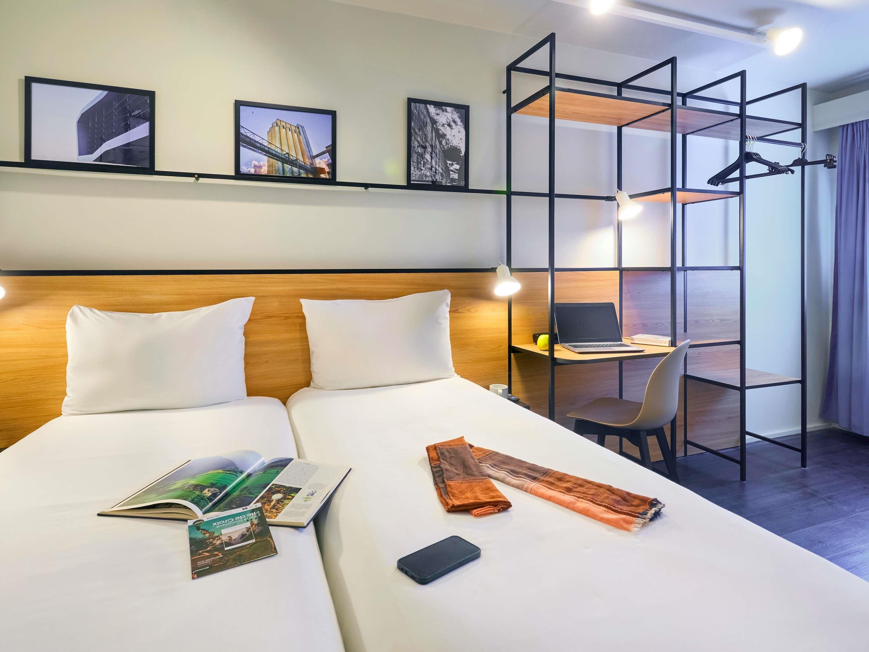 A hotel room with a bed, a desk with a laptop, and a chair. There are pictures on the wall and a shelf with a lamp.