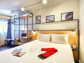 Hotelkamer met een stapelbed, bureau met een laptop, een stoel, bed met kussens, nachtkastje met een lamp, en ingelijste foto’s aan de muur.