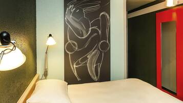 A hotel room with a bed, a wall-mounted lamp, and a red door.