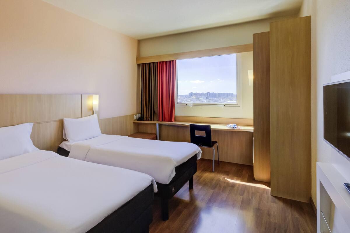 A hotel room with two beds, a desk, a chair, a TV, and a window with a city view.