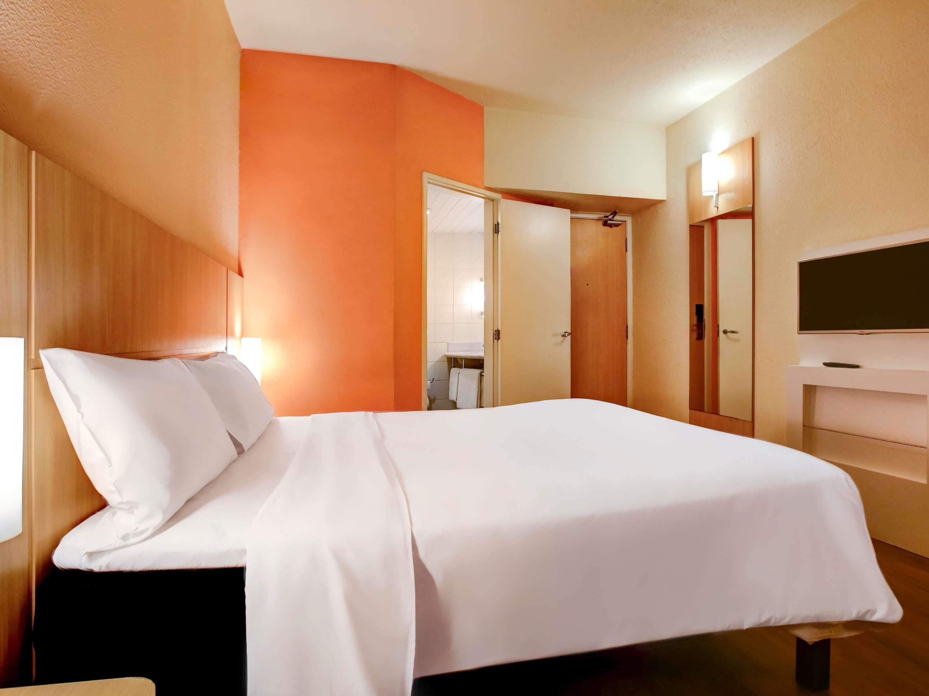 Habitación de hotel con cama, televisor, armario y baño visible a través de una puerta abierta.