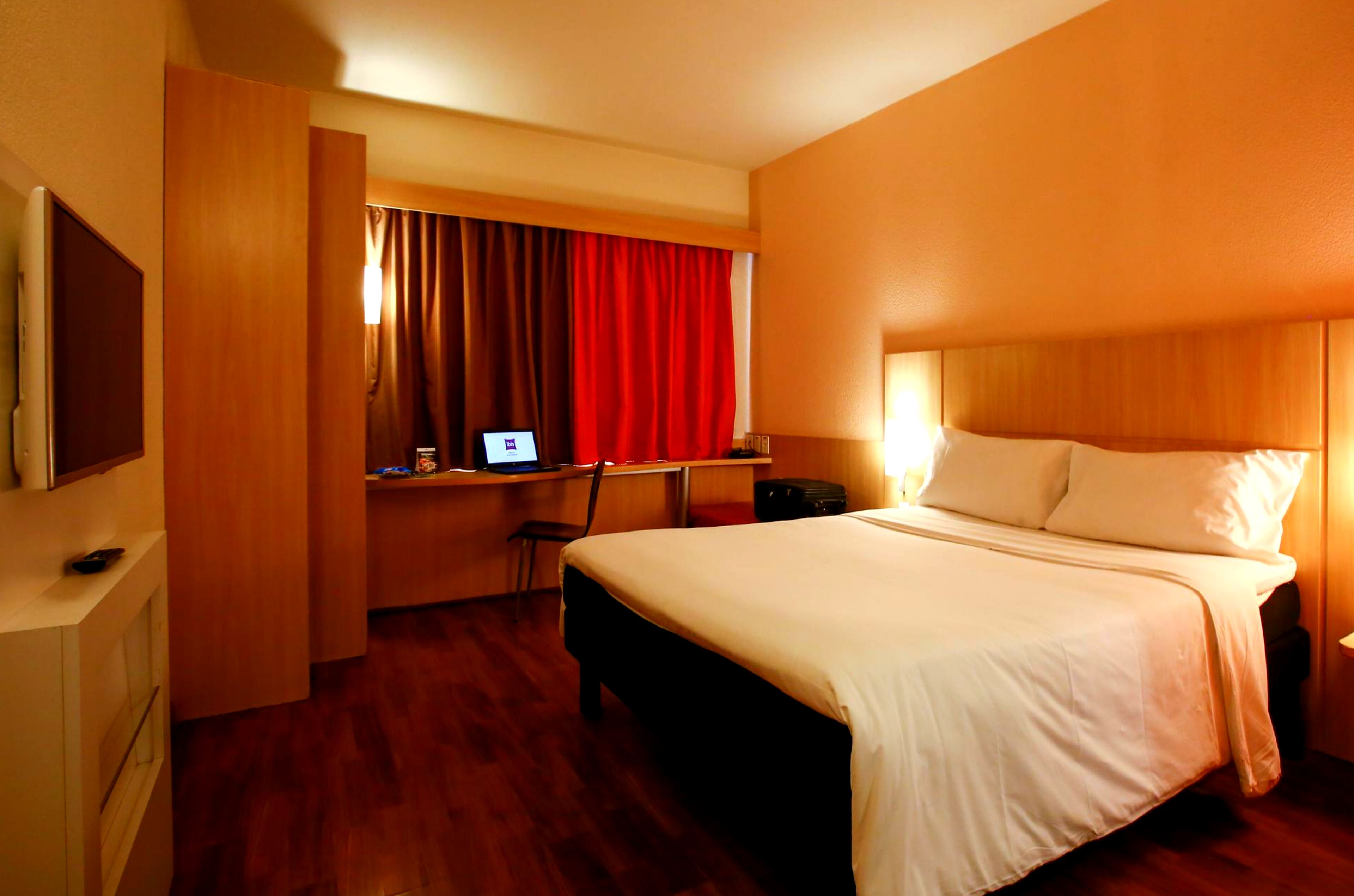 Una habitación de hotel con cama, escritorio, computadora, silla y televisión en la pared.