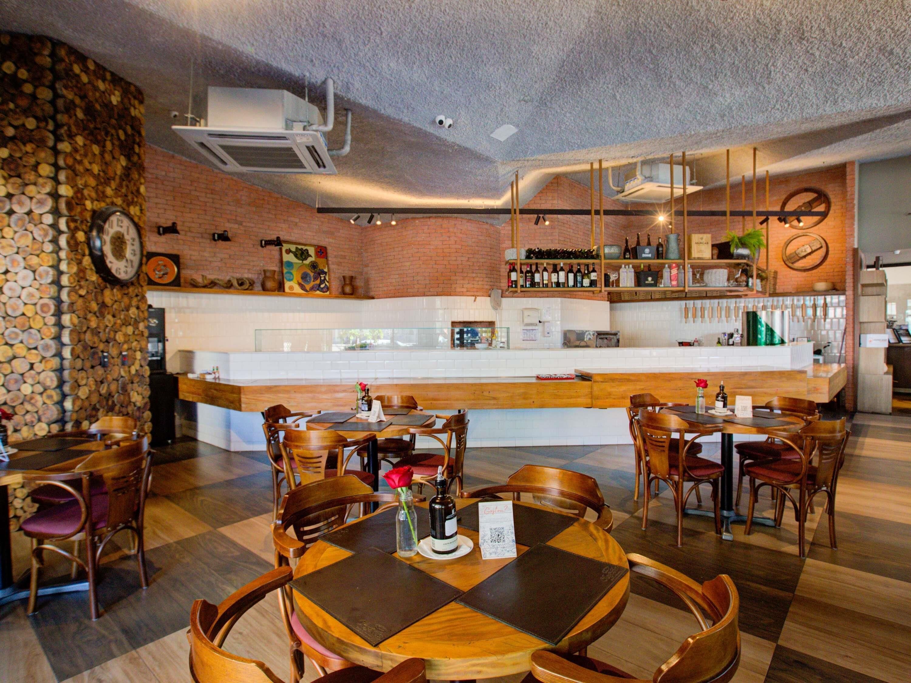 Un restaurante con pared de ladrillo, mesas y sillas de madera, una zona de bar con botellas y un reloj en la pared.