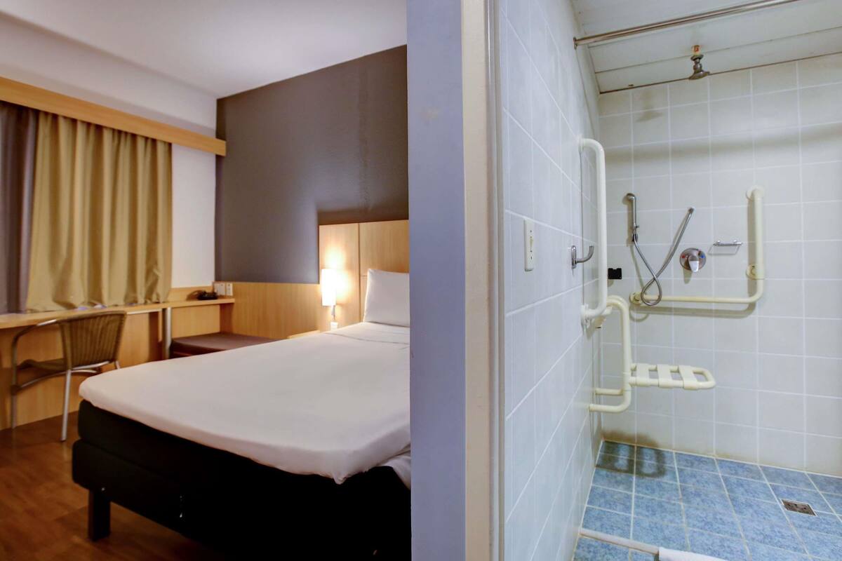 A hotel room with a bed, a desk, and a shower area with grab bars.