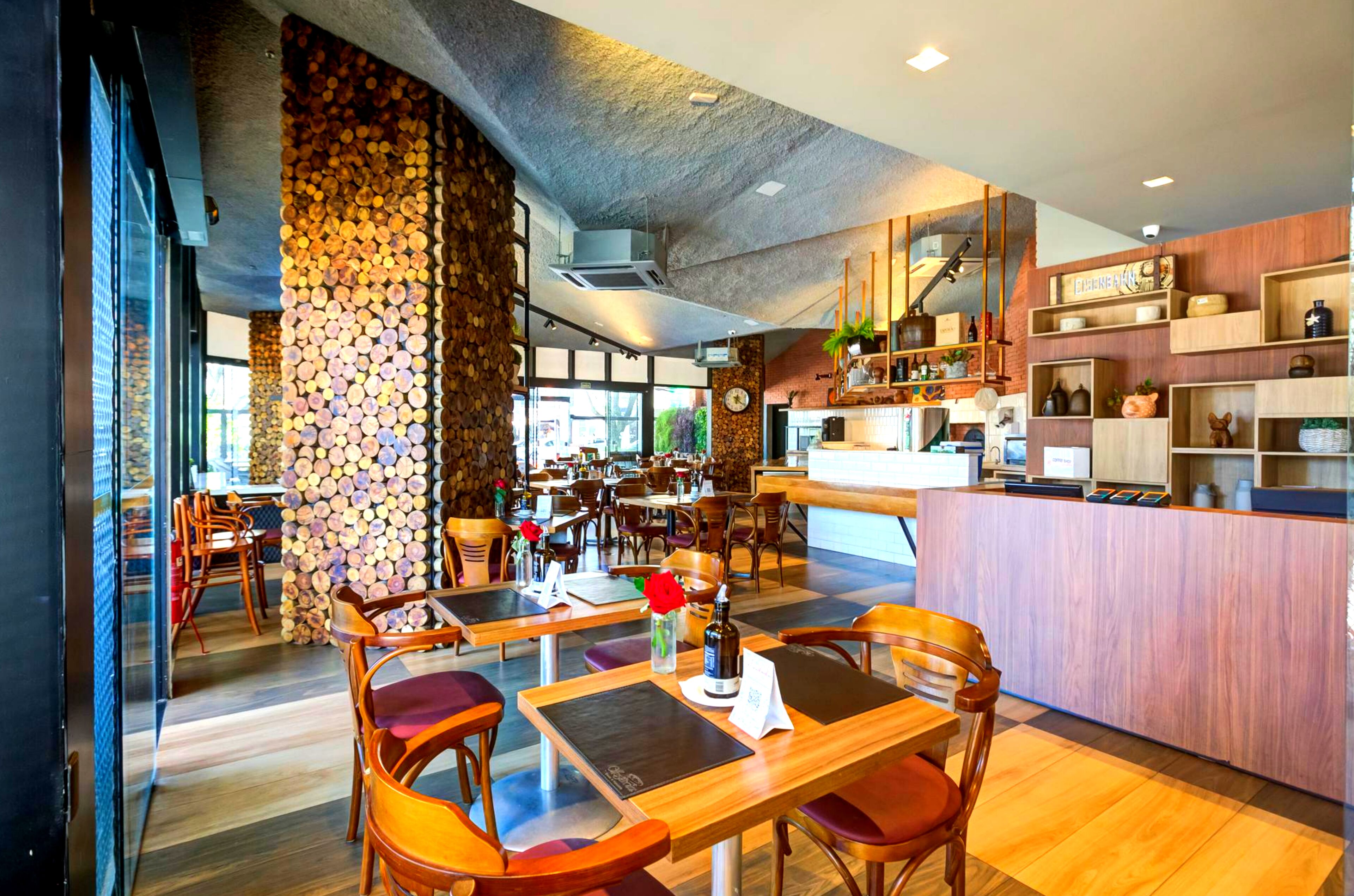 Restaurante moderno com mobiliário de madeira, parede de pedra e área de cozinha visível ao fundo.