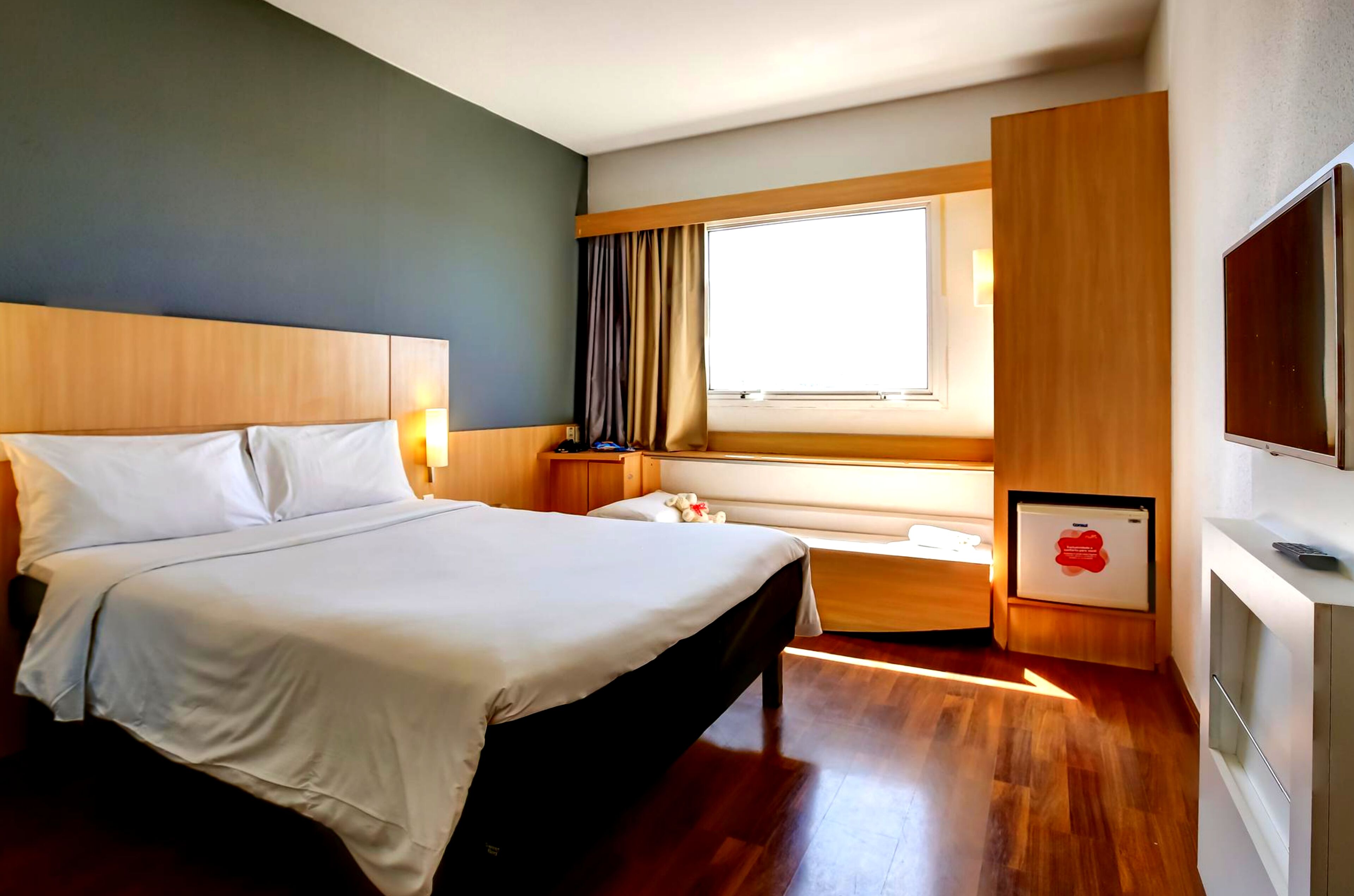A hotel room with a large bed, a bedside table, a TV, and a window with curtains.