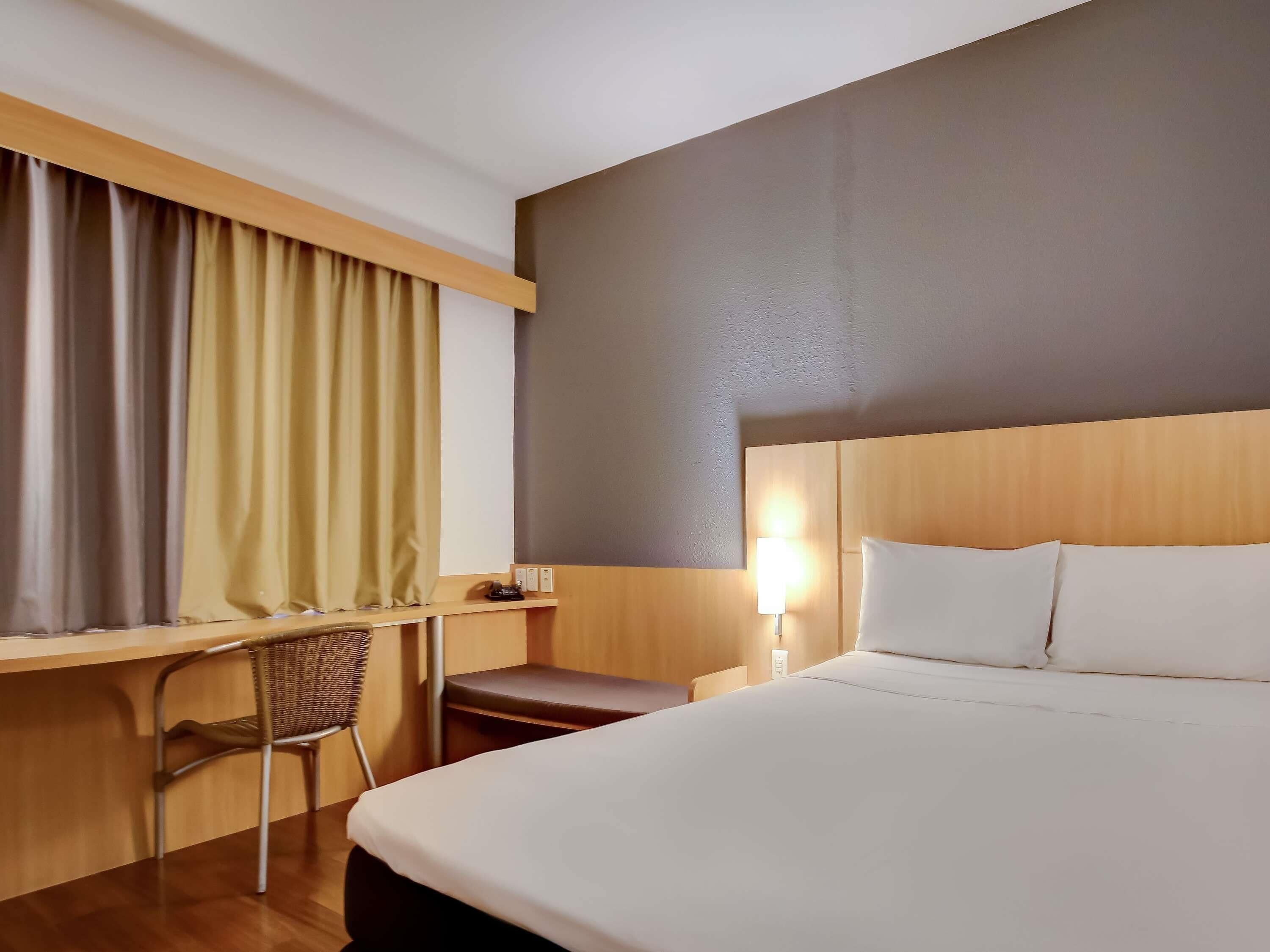 A hotel room with a bed, desk, chair, and a nightstand with a lamp.
