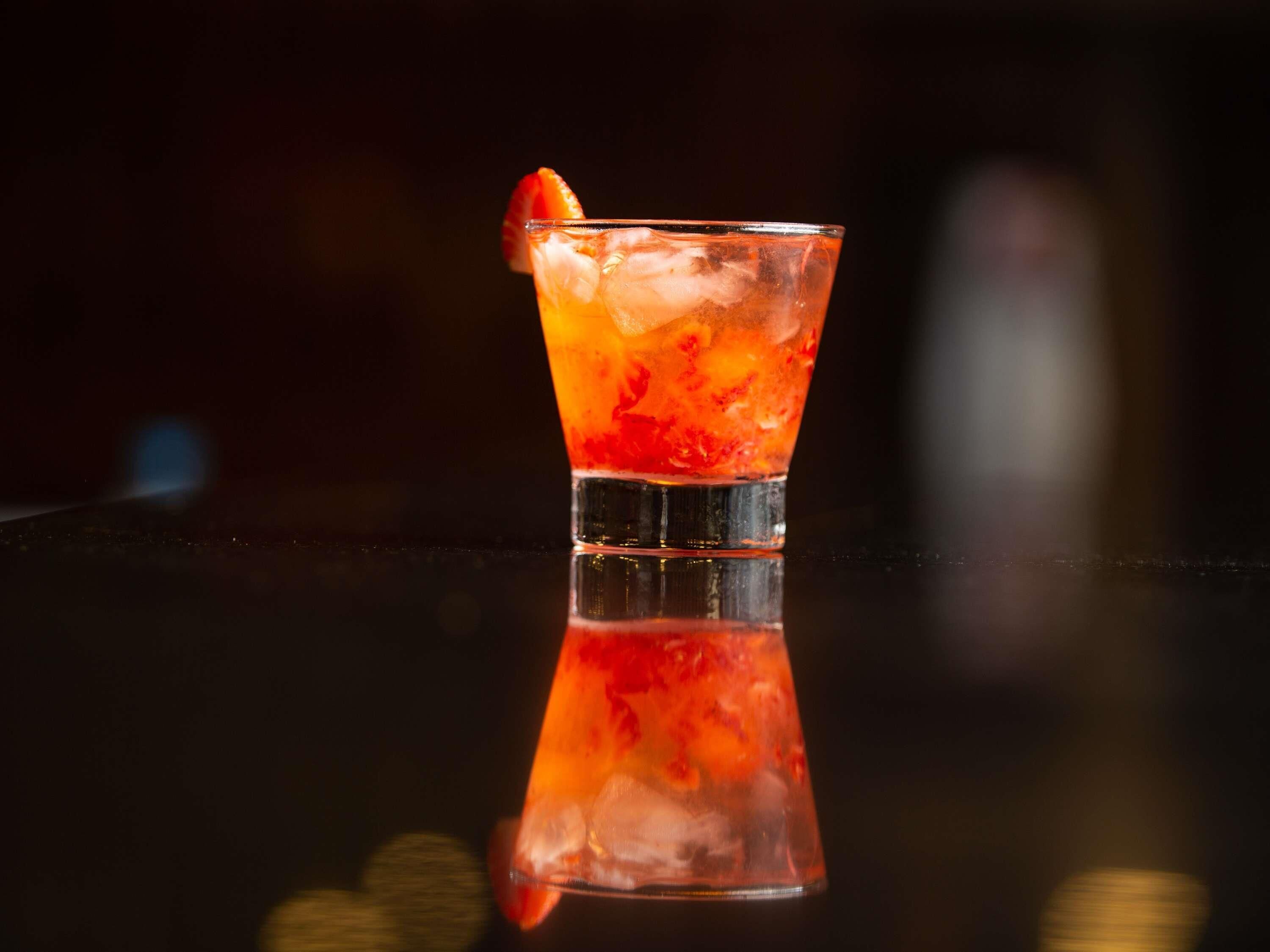 A glass of red beverage with ice cubes and a strawberry garnish on a reflective surface.