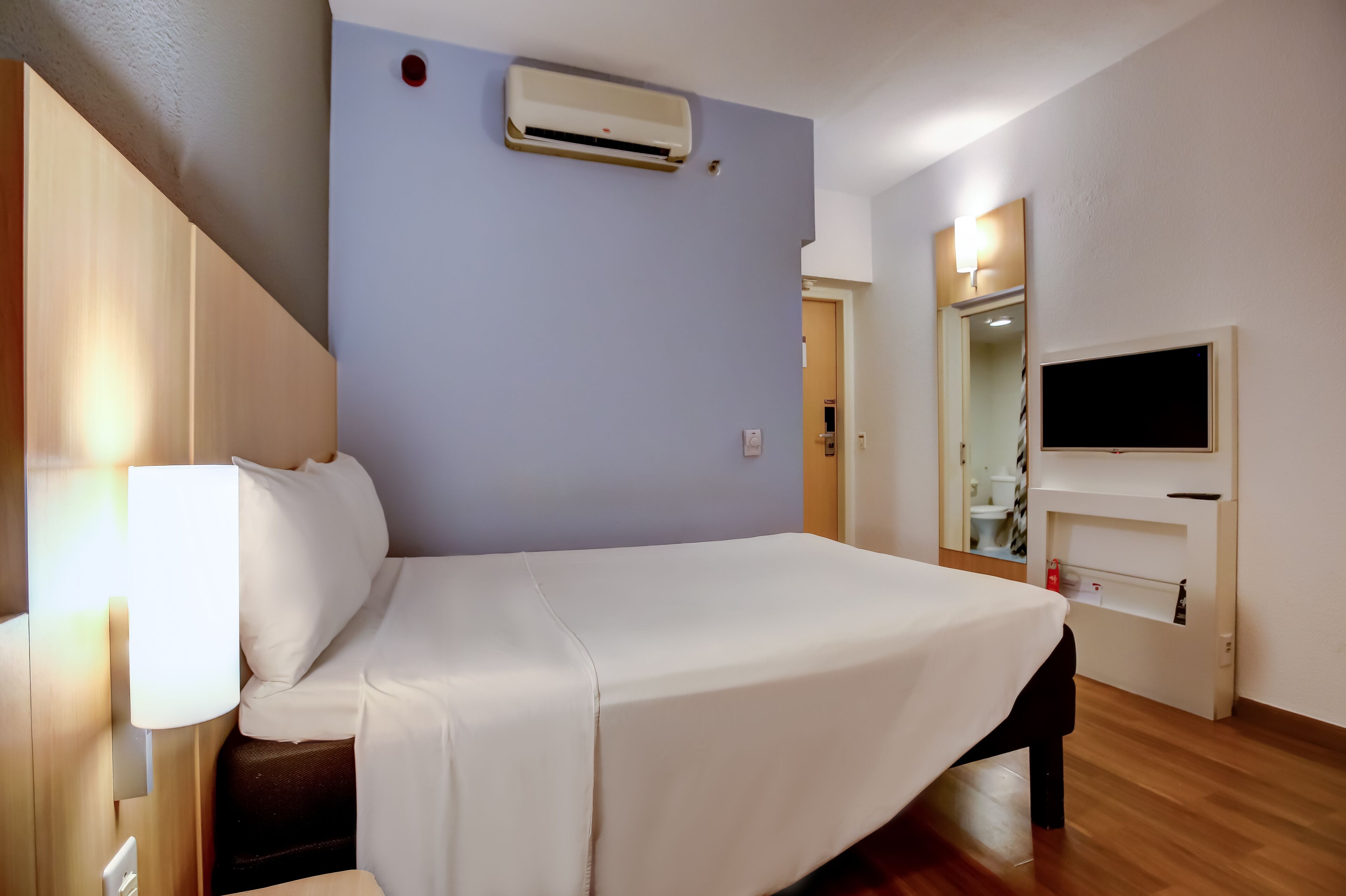 A hotel room with a bed, bedside lamp, air conditioning unit, TV, and a mirror.