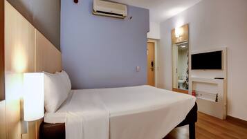 A hotel room with a bed, bedside lamp, air conditioning unit, TV, and a mirror.