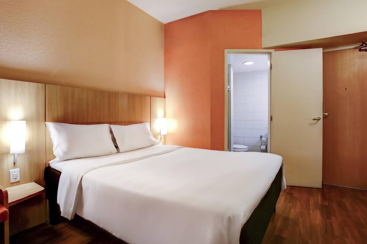 A hotel room with a wooden headboard, a white bedspread, a nightstand with a lamp, and a wooden floor.