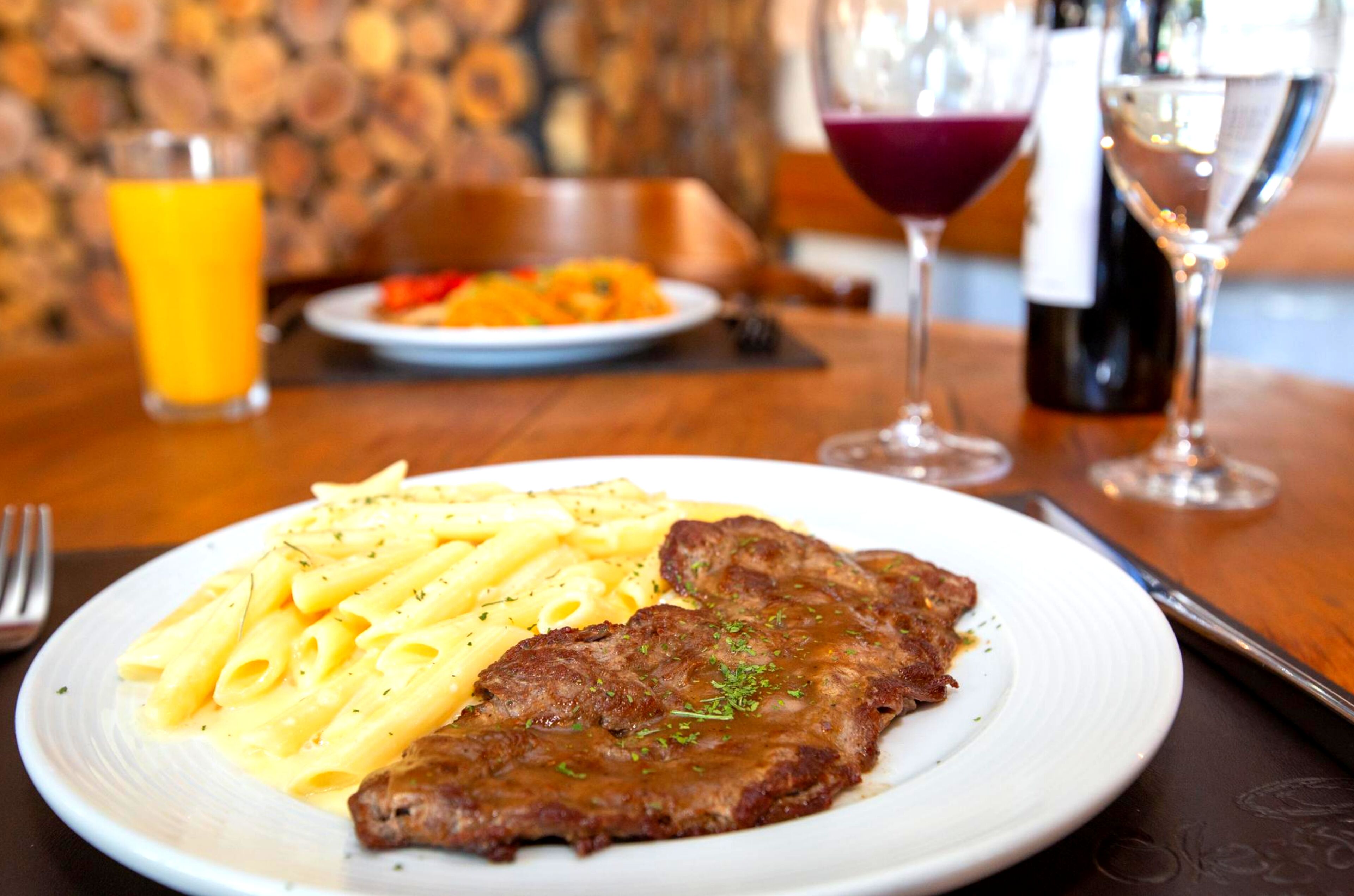 Un plato de pasta y filete sobre una mesa, junto a un vaso de jugo de naranja y una botella de vino.