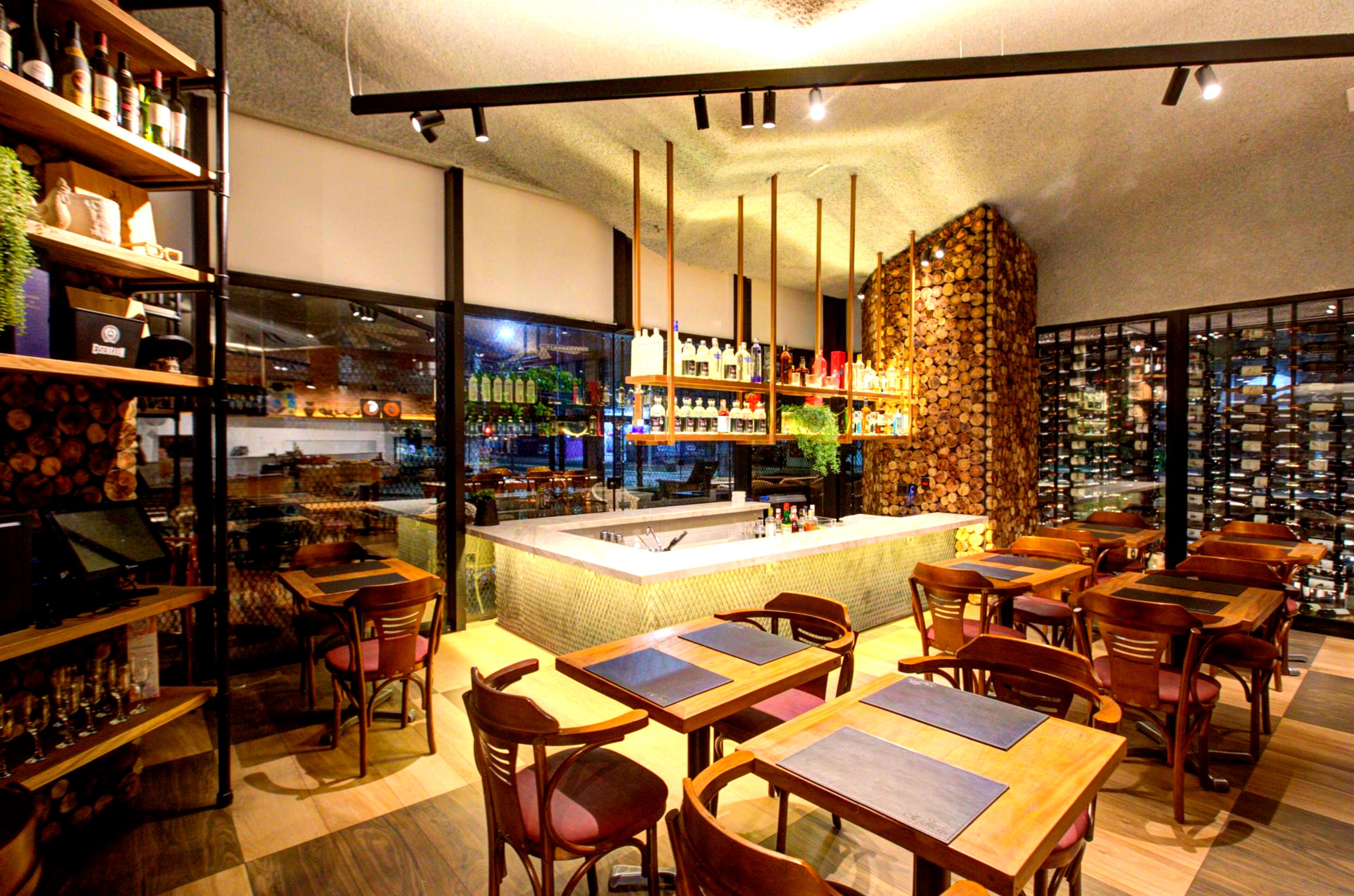 A restaurant with wooden tables and chairs, a bar area with shelves of bottles, and a large window with a view of the city.