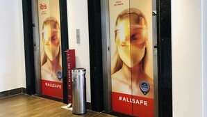 A hallway with two elevators, each with a large advertisement for 'ibis' featuring a woman and the hashtag '#ALLSAFE'. - ibis Vitoria Praia Do Canto (Vitoria)