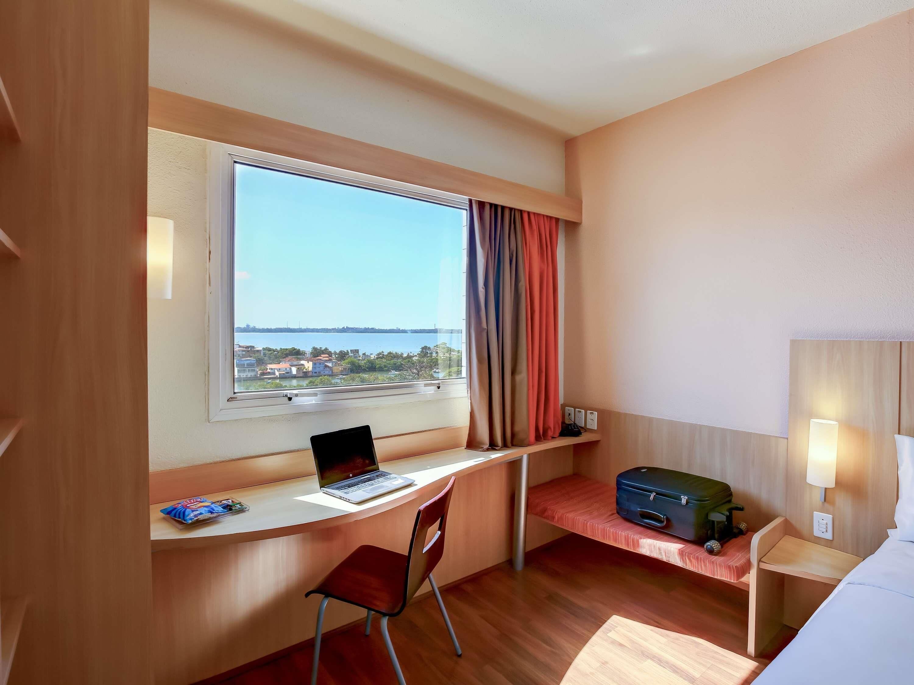 Quarto de hotel com escrivaninha, cadeira, laptop, mala e cama.