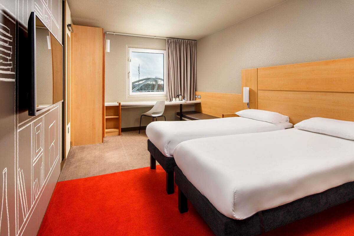 A hotel room with two beds, a desk, a chair, and a TV.