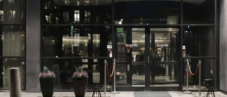 L’entrée d’un hôtel, avec des portes en verre, une barrière faite d’une corde rouge et des plantes en pot disposées sur le trottoir.