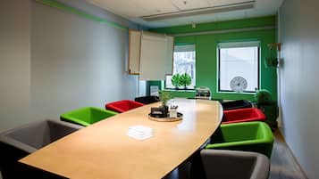 Un salón de conferencias con una larga mesa, sillas verdes y rojas, un reloj y una ventana con persianas.