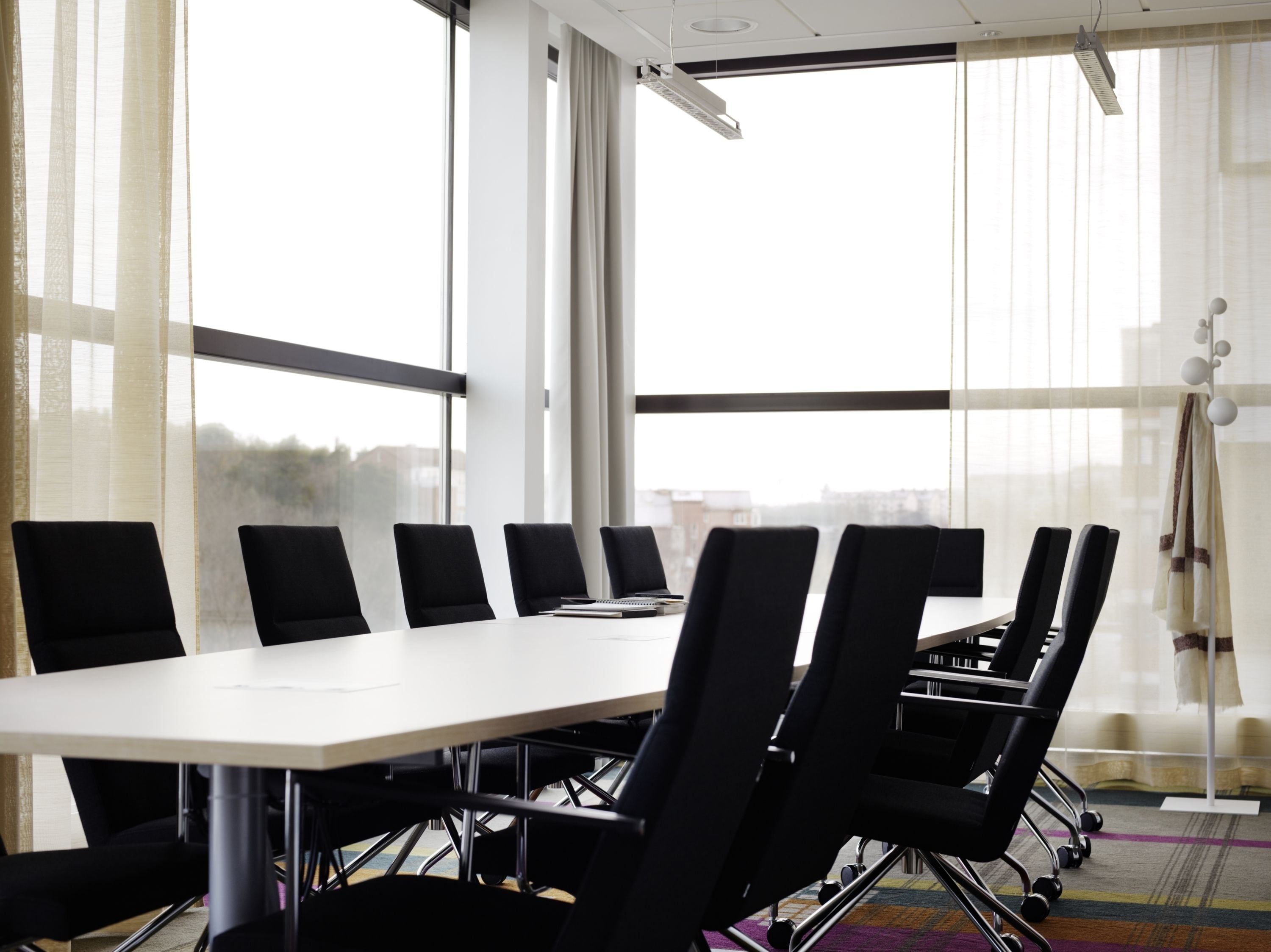 A conference room with a long table and black chairs, large windows with sheer curtains, and a city view.