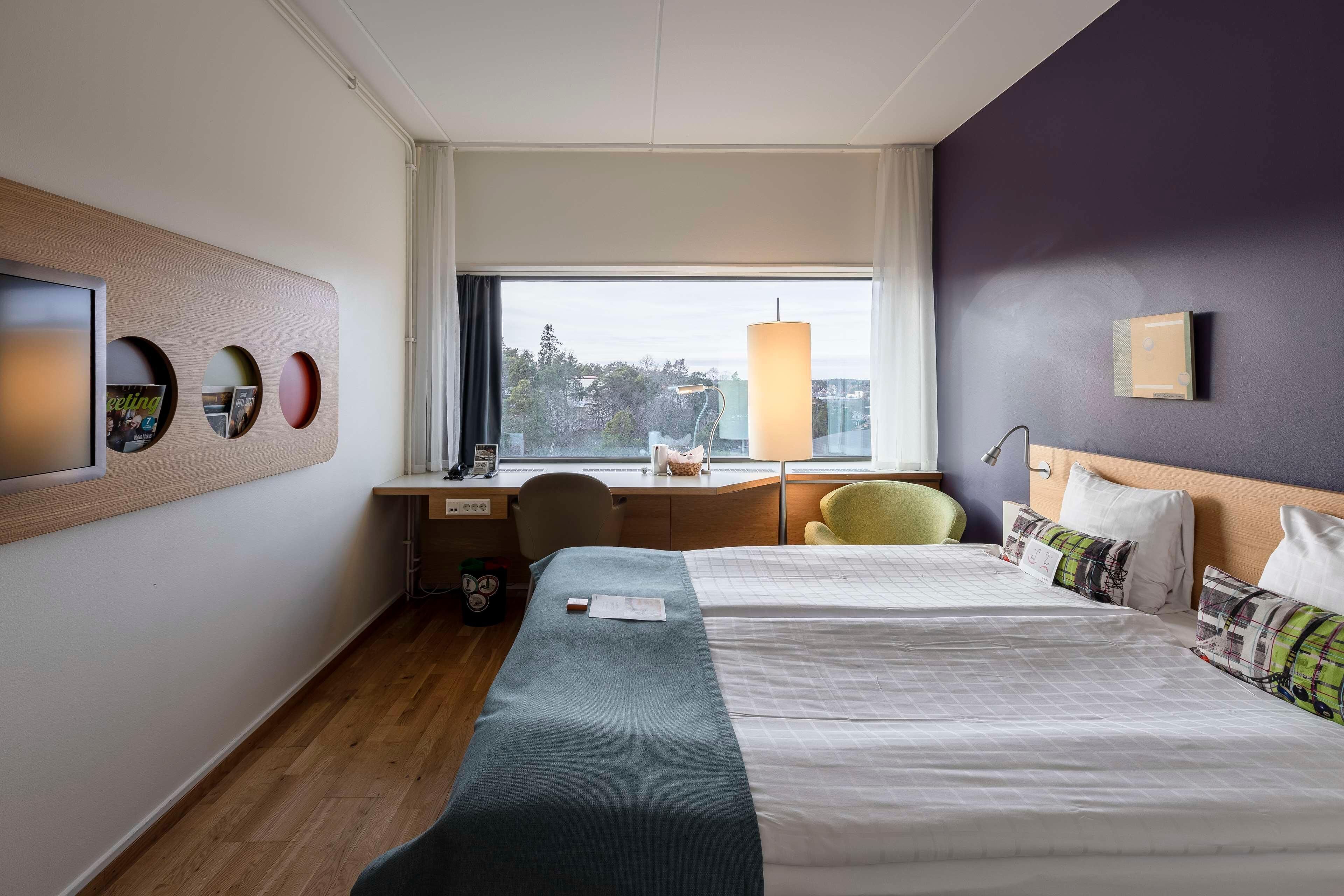Habitación de hotel con dos camas, un escritorio, una silla, una lámpara y ventana con vista a árboles y edificios.
