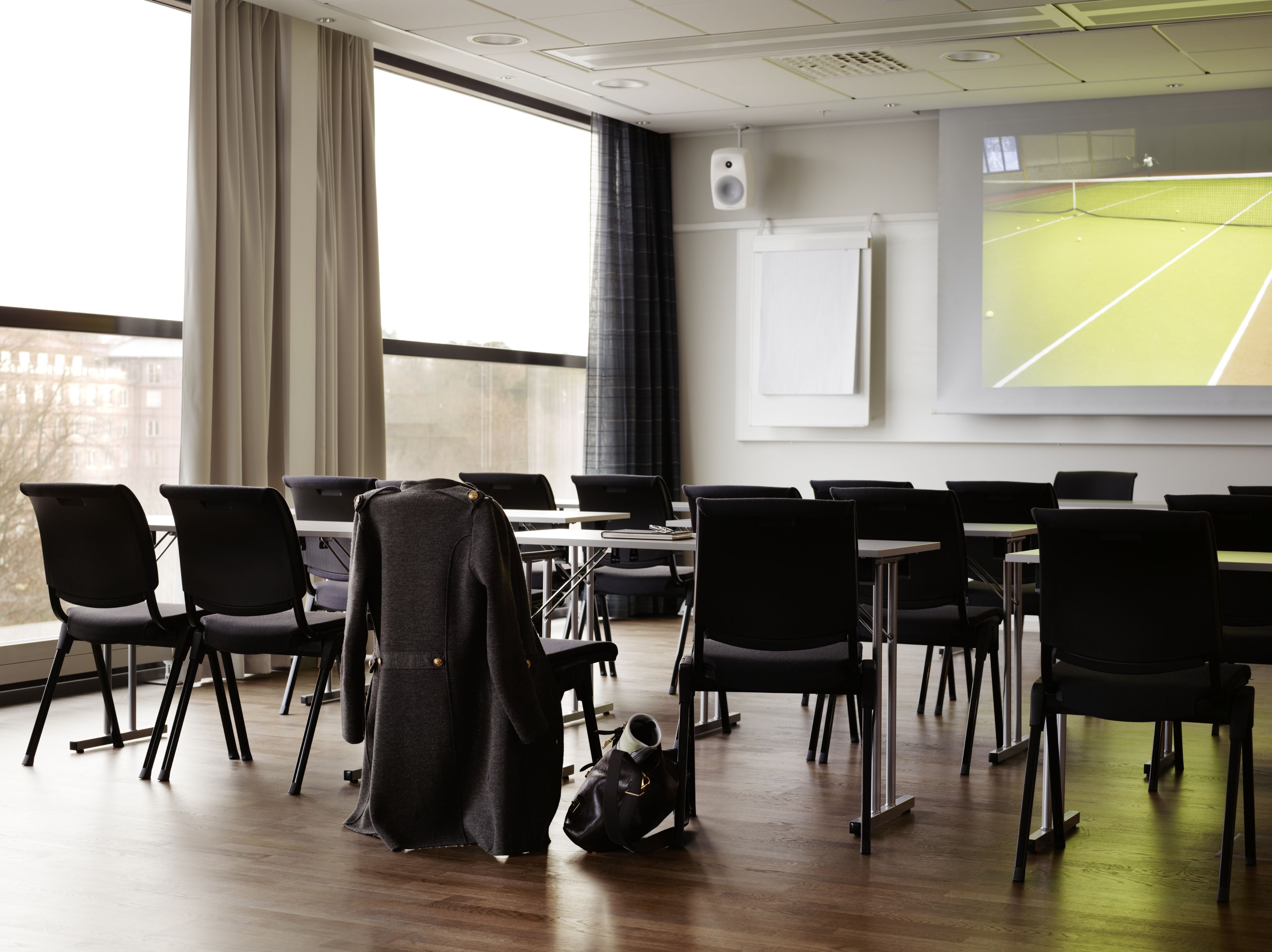 Un salón de conferencias con una pantalla grande que muestra una cancha de tenis, sillas negras, una mesa y un abrigo colgado en una silla.