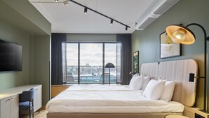 A modern hotel room with a large bed, a desk, and a view of the city through large windows. - Scandic Alvik (Bromma)